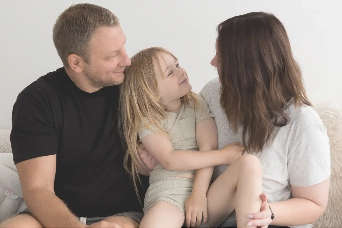 Our founding family sitting together on a light-colored couch. The father on the left wears a black t-shirt and looks at his daughter, who is in the center wearing a short set and looking up at her mother on the right, who wears a white t-shirt.
