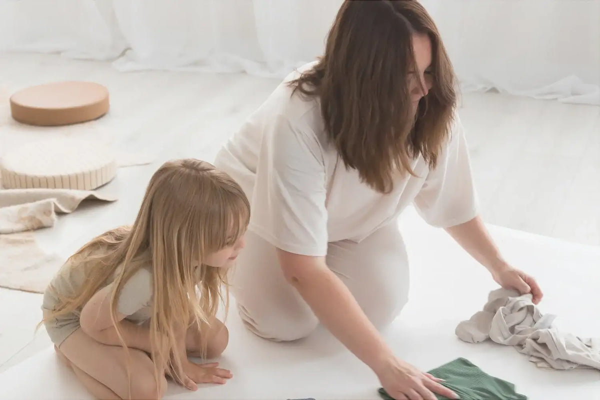 Our founder and her daughter are kneeling on a white floor in a bright, minimalist room. Our founder is sorting through light-coloured clothing, while the daughter looks on.
