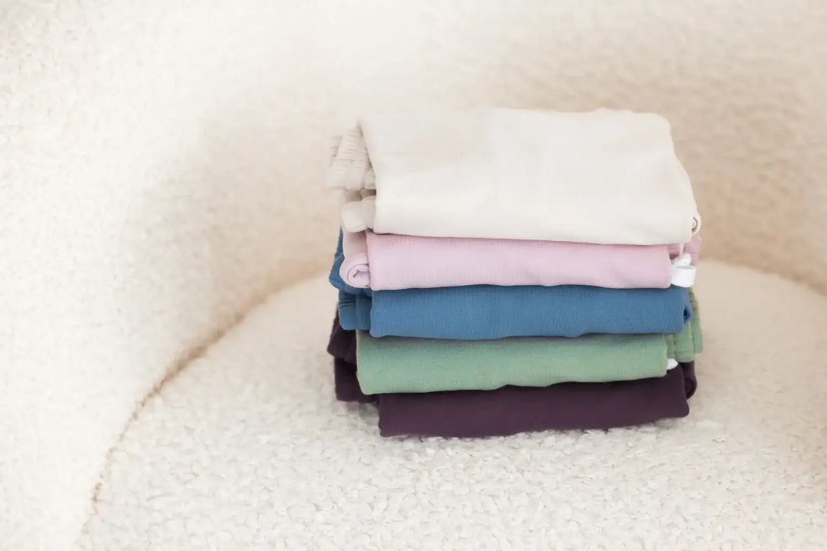 A stack of five folded organic cotton jogger sets in different colours, including beige, pink, blue, sage green, and dark purple, arranged on a textured cream surface.