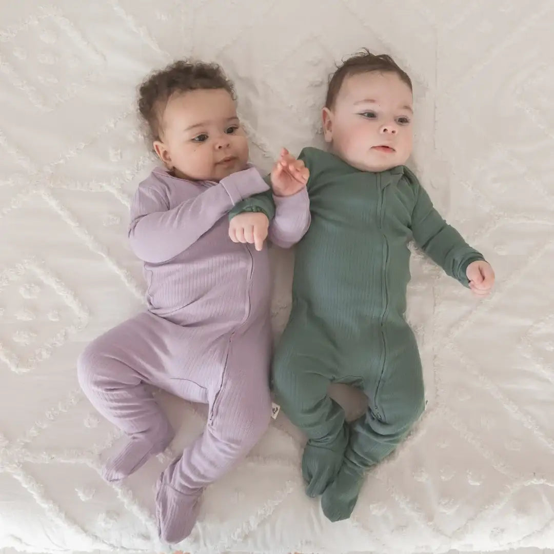 Two babies lying side by side on a white textured blanket, wearing ribbed footed sleeper onesies in Lavender Blush and Pine.