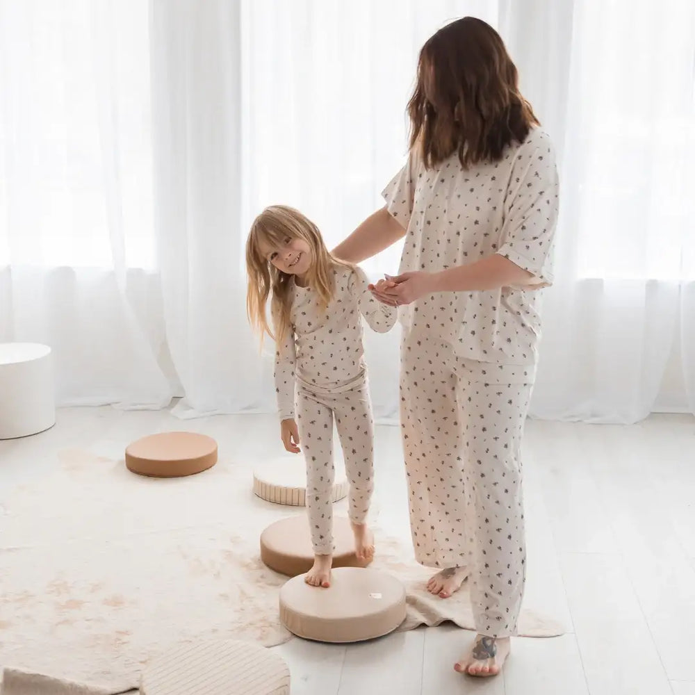A woman and a young girl wearing matching pajamas with small floral print are barefoot on round wooden cushions in a bright, airy room with white curtains.