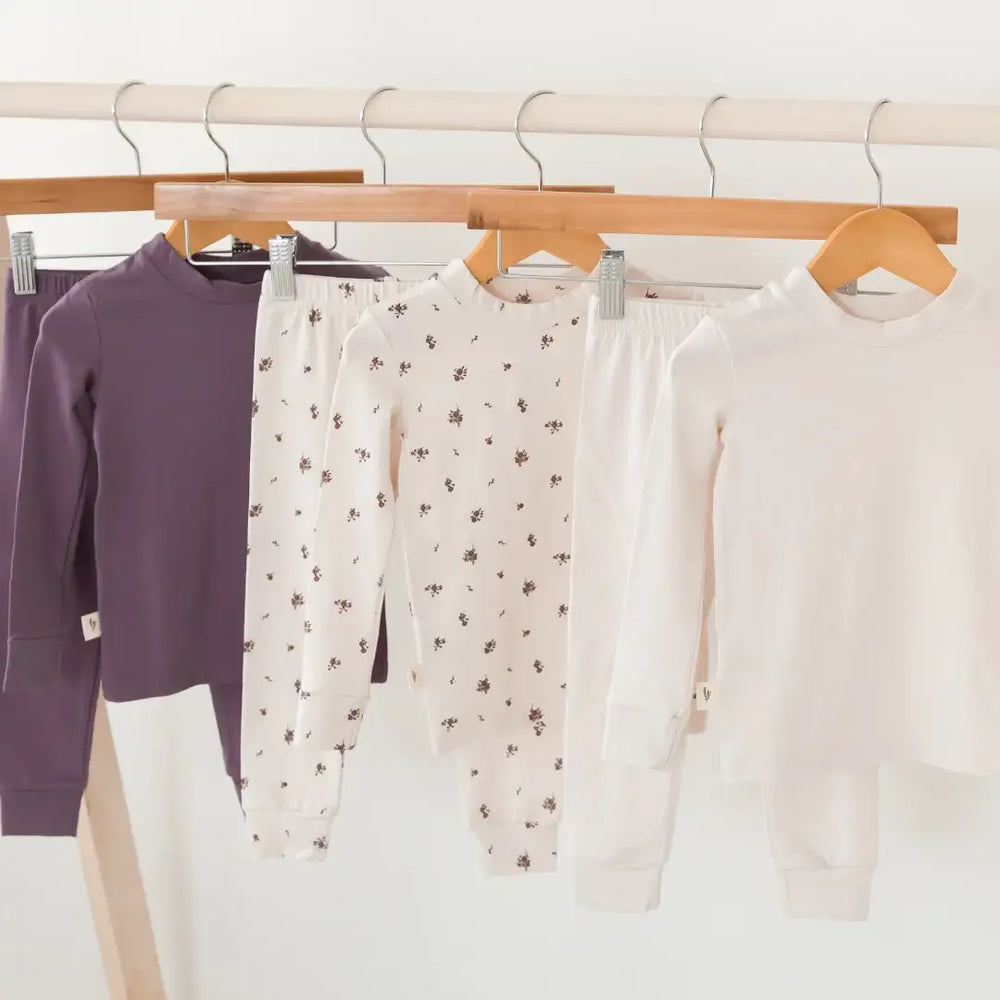 Three children's long-sleeve pajama sets hanging on wooden hangers from a clothing rack. The sets are in Wild Mulberry, Birch, and Birch Floral, by Milana Label.