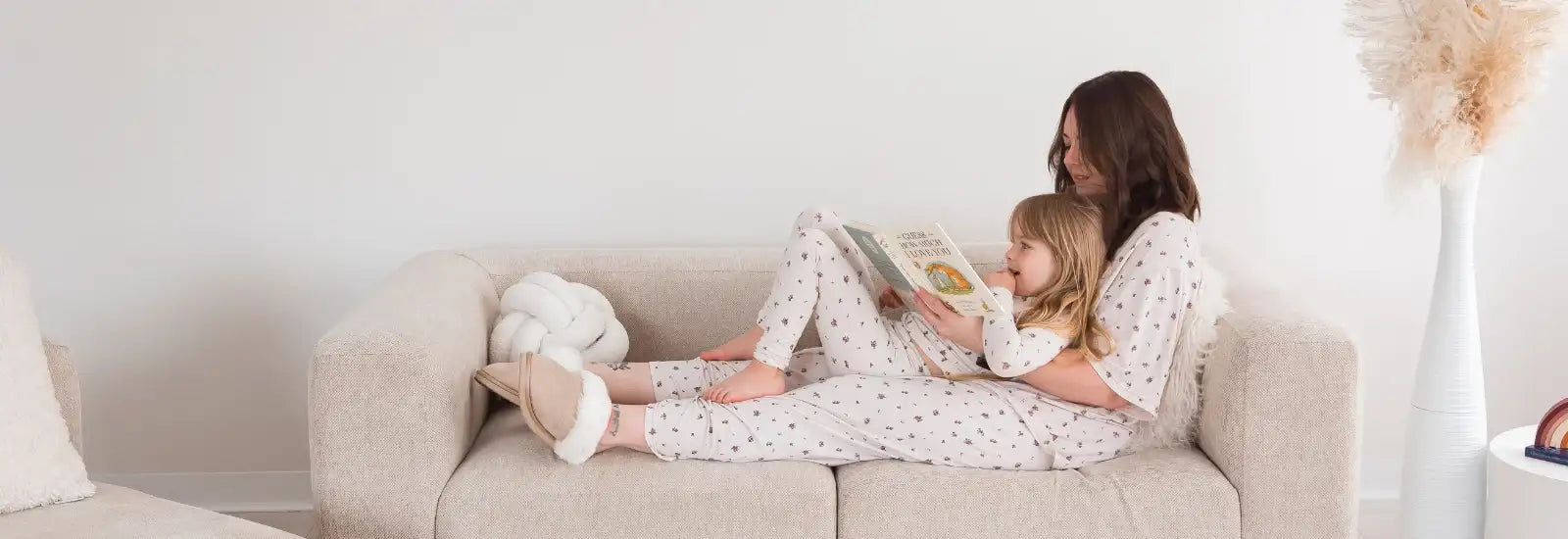 A woman and a young girl sitting together on a light beige sofa, both wearing matching Milana pajamas with a small floral pattern. The woman is holding an open book, and a white plush pillow rests beside them on the couch in a bright, minimal living room.