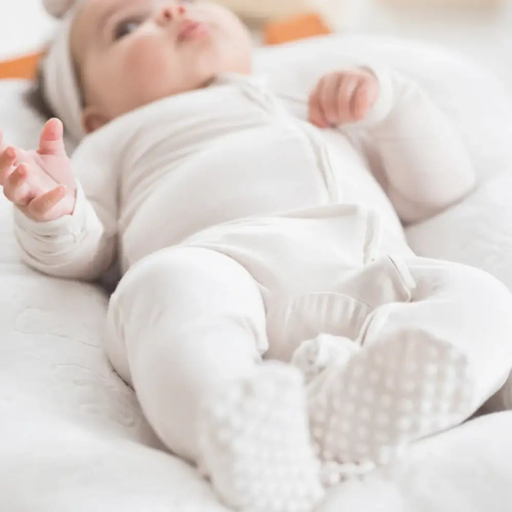 A baby lying on a white quilted lounger, wearing a greige long-sleeve footed onesie and matching headband.