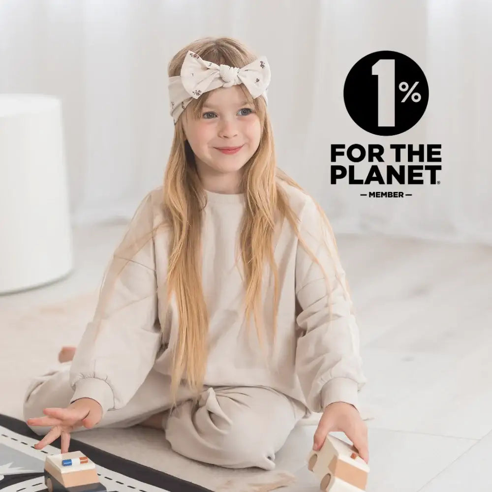 A young girl with long blonde hair sits cross-legged on a light-colored floor, wearing a beige sweatshirt and matching pants with a bow headband. 1% for the Planet member is overlayed.