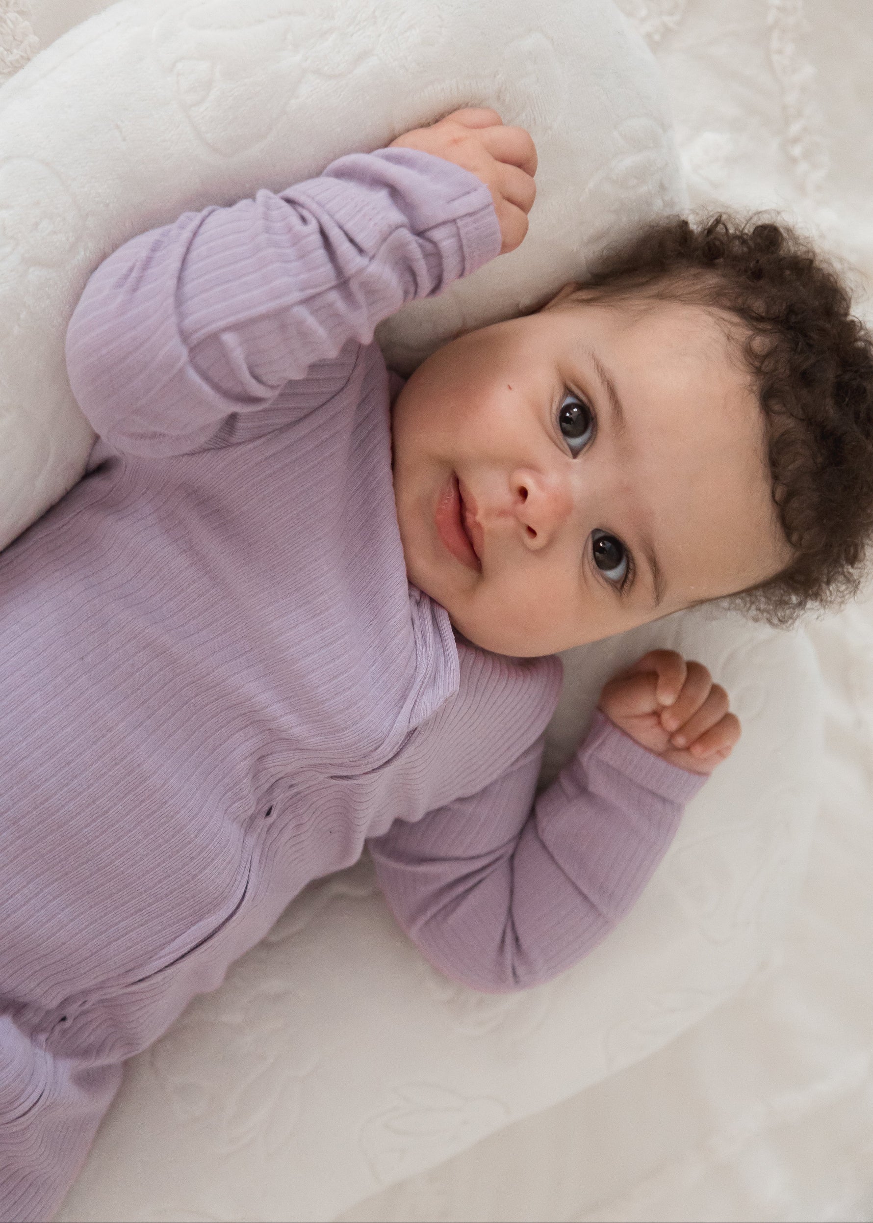 Angled close-up shot of a baby looking directly at the camera while wearing the Lavender Blush ribbed baby sleeper PJ.