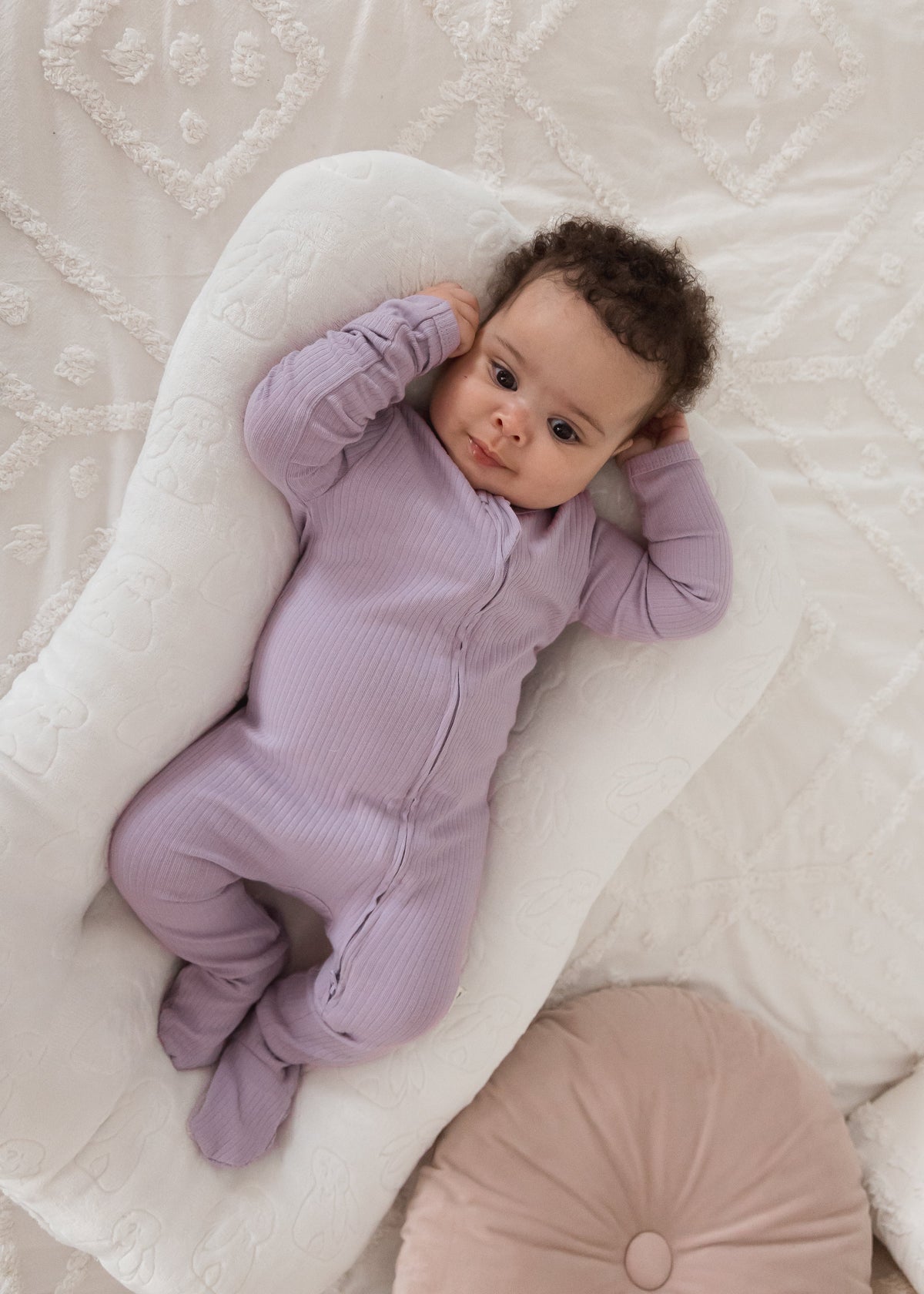 Overhead view of a baby lounging in a white baby nest wearing the Lavender Blush ribbed organic cotton footie sleeper next to a pink decorative cushion.