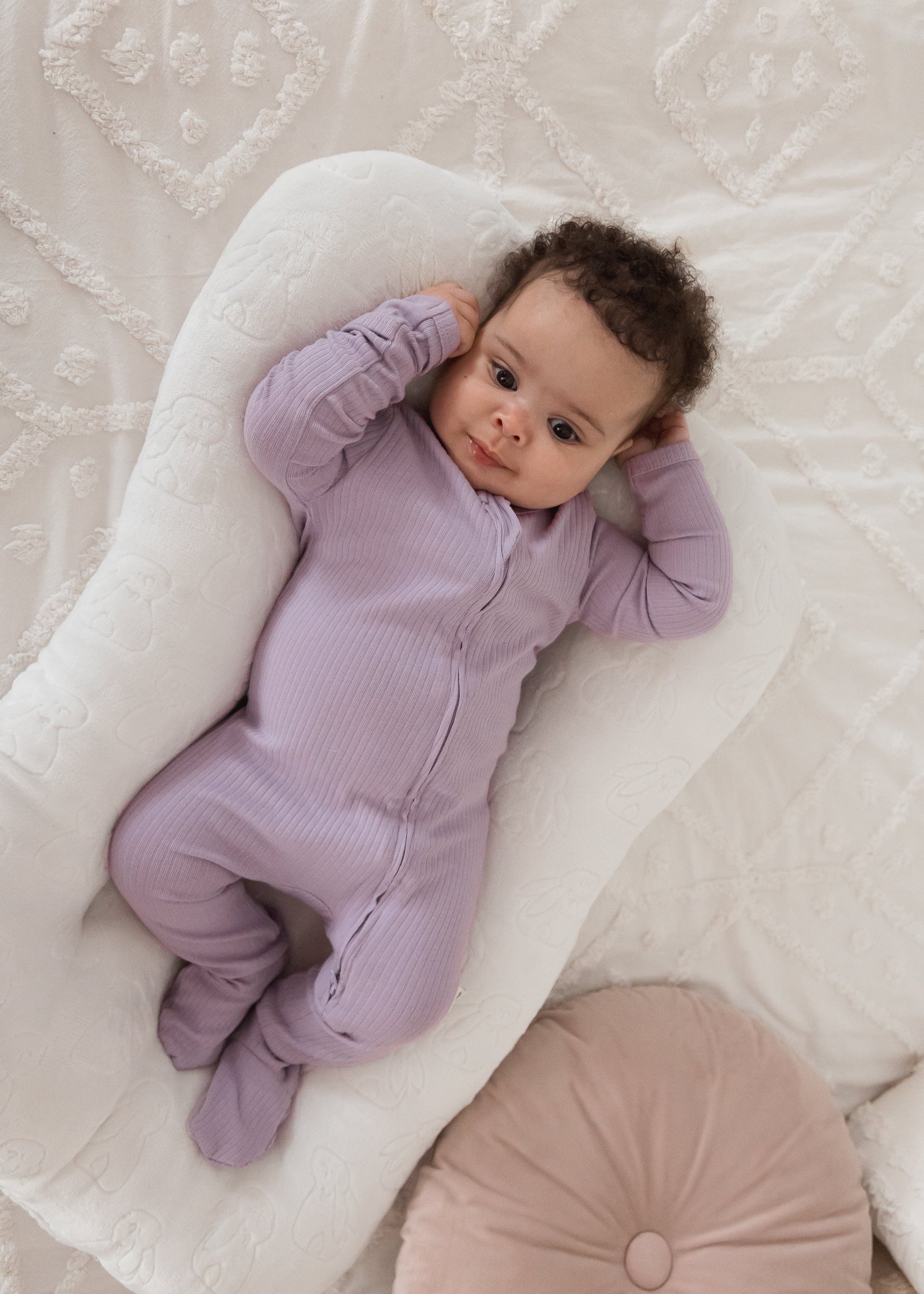 Overhead view of a baby lounging in a white baby nest wearing the Lavender Blush ribbed organic cotton footie sleeper next to a pink decorative cushion.