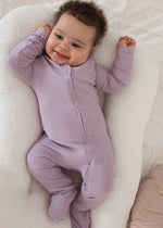 A happy baby smiling and stretching while wearing the Milana Label Lavender Blush organic cotton ribbed footie sleeper in a cream baby lounger.