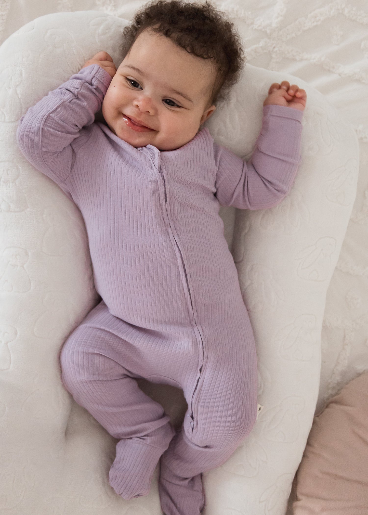 A happy baby smiling and stretching while wearing the Milana Label Lavender Blush organic cotton ribbed footie sleeper in a cream baby lounger.