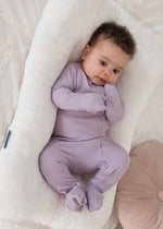 Full-length view of a baby looking at the camera while wearing the Milana Label Lavender Blush ribbed footie sleeper in a plush white lounger.