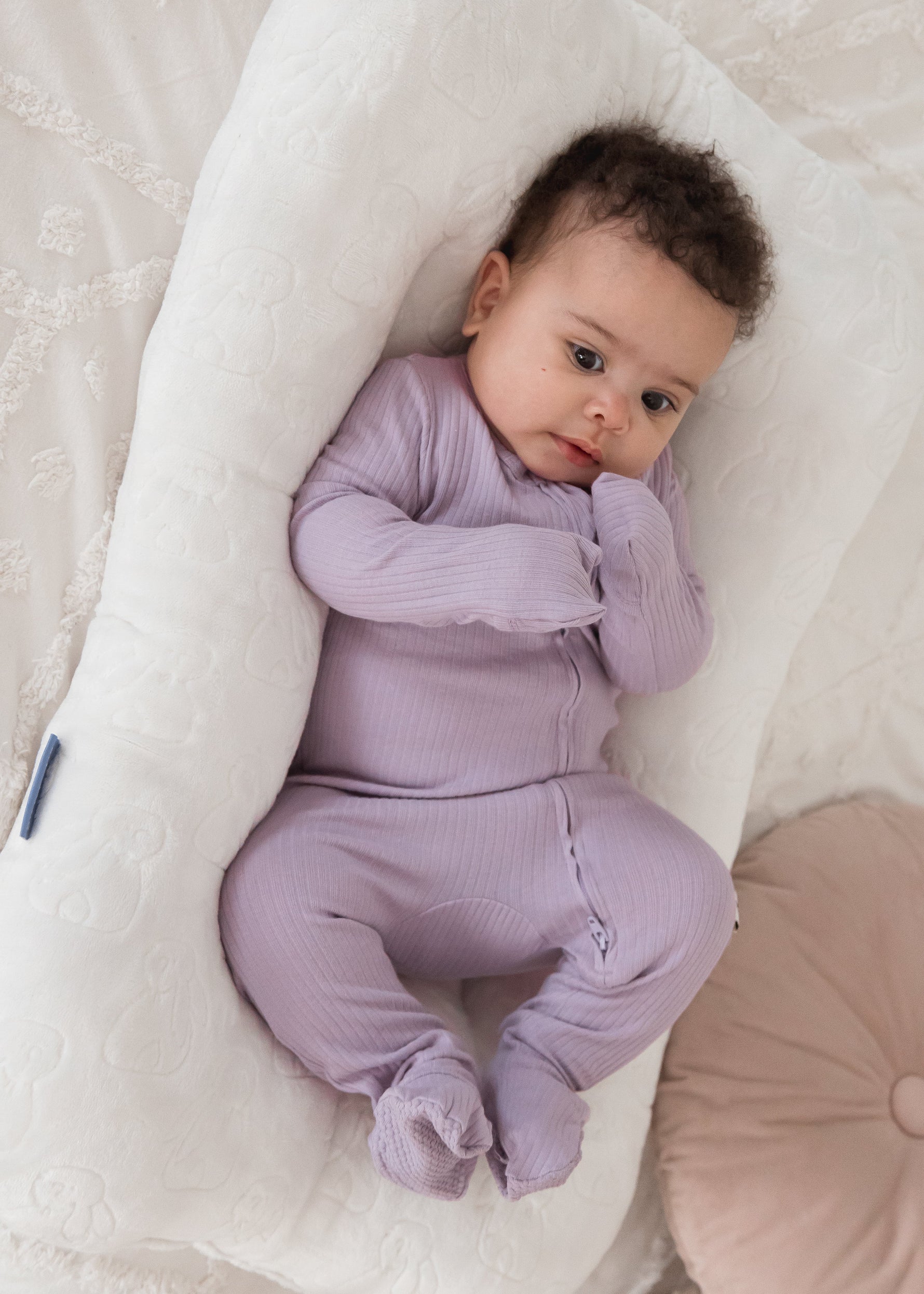 Full-length view of a baby looking at the camera while wearing the Milana Label Lavender Blush ribbed footie sleeper in a plush white lounger.