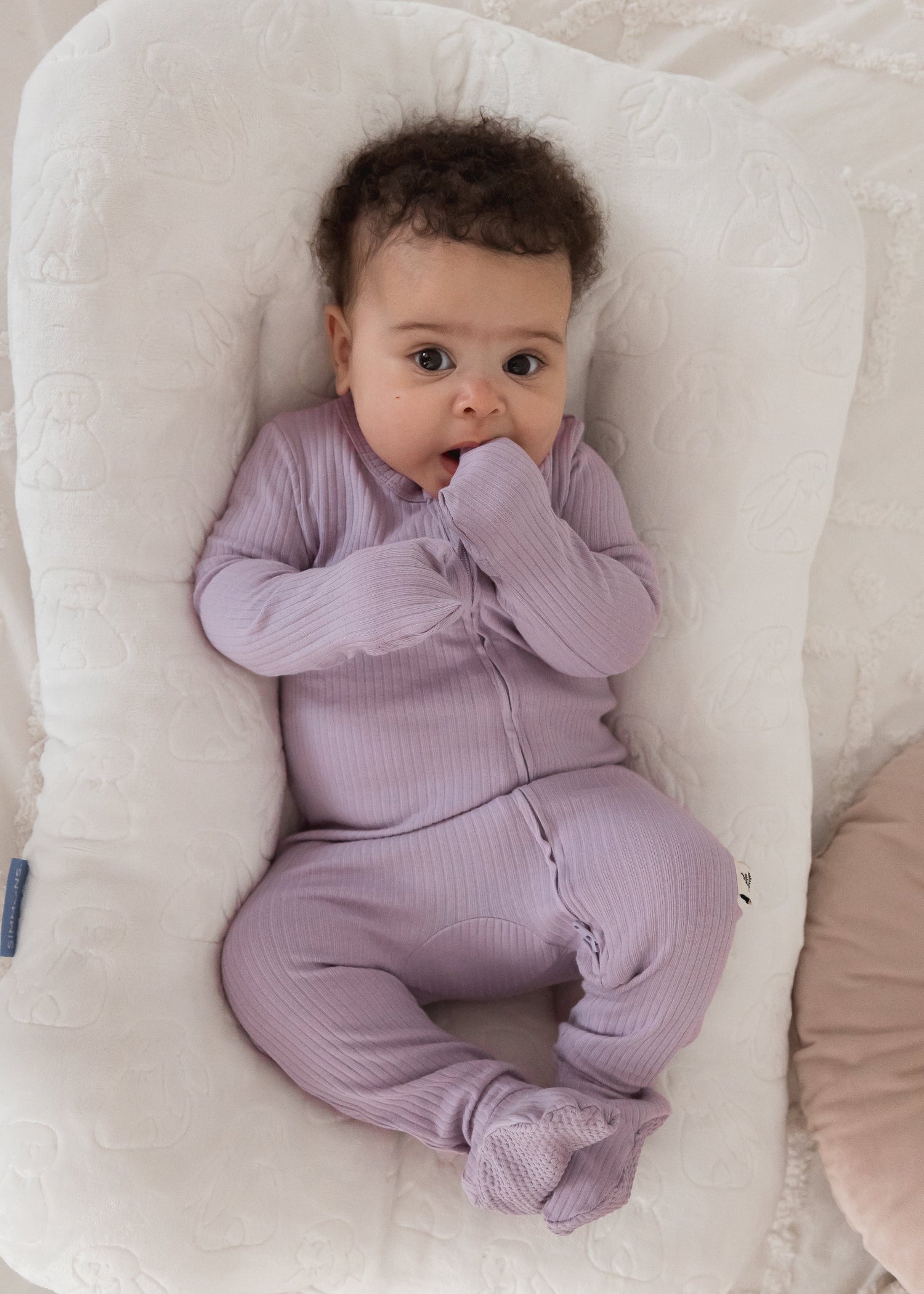 A baby wearing the Lavender Blush ribbed organic cotton footie sleeper while lounging comfortably in a plush cream-colored baby nest.