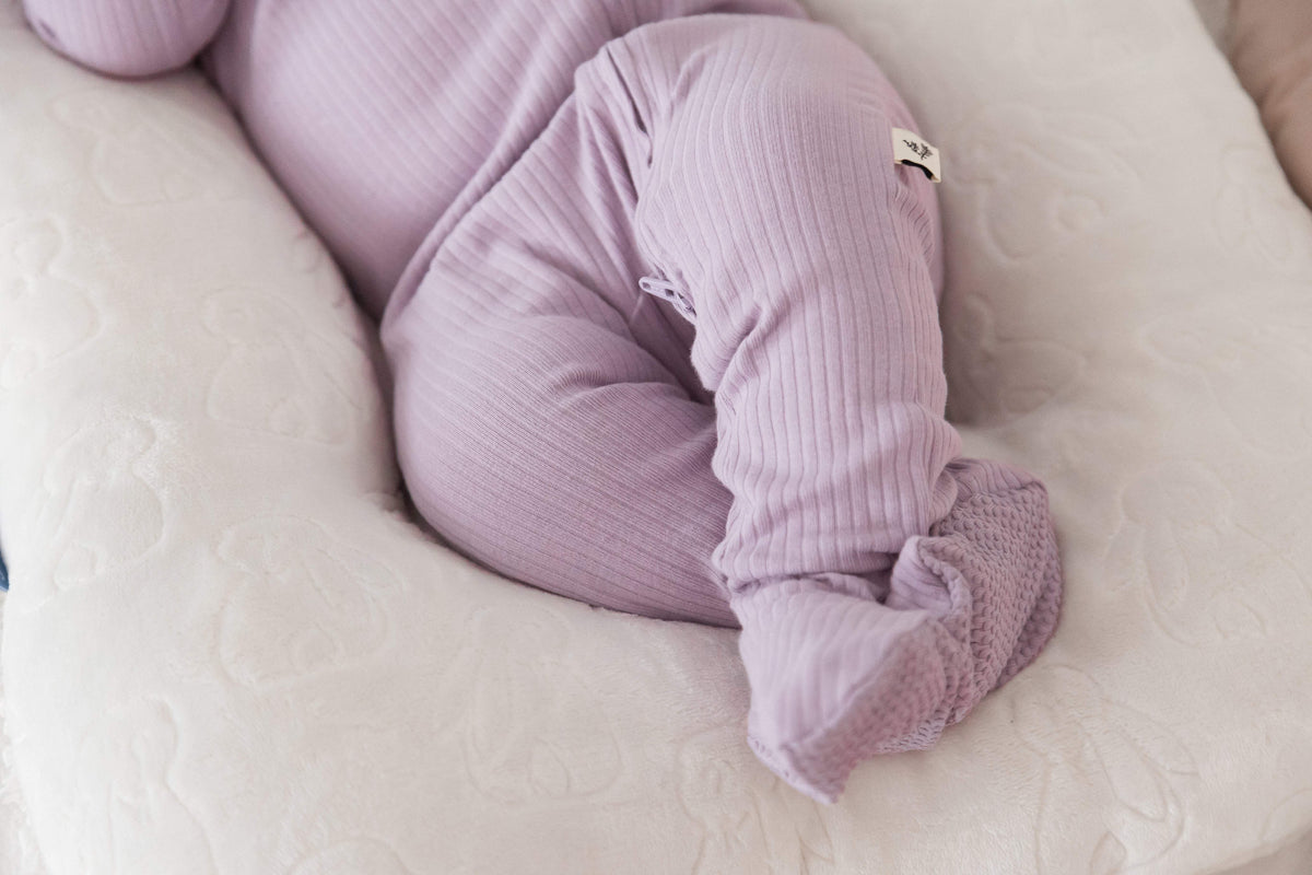 A close-up of a baby wearing a ribbed organic sleeper in the colour Lavender Blush, curled up on a textured white-coloured lounger.