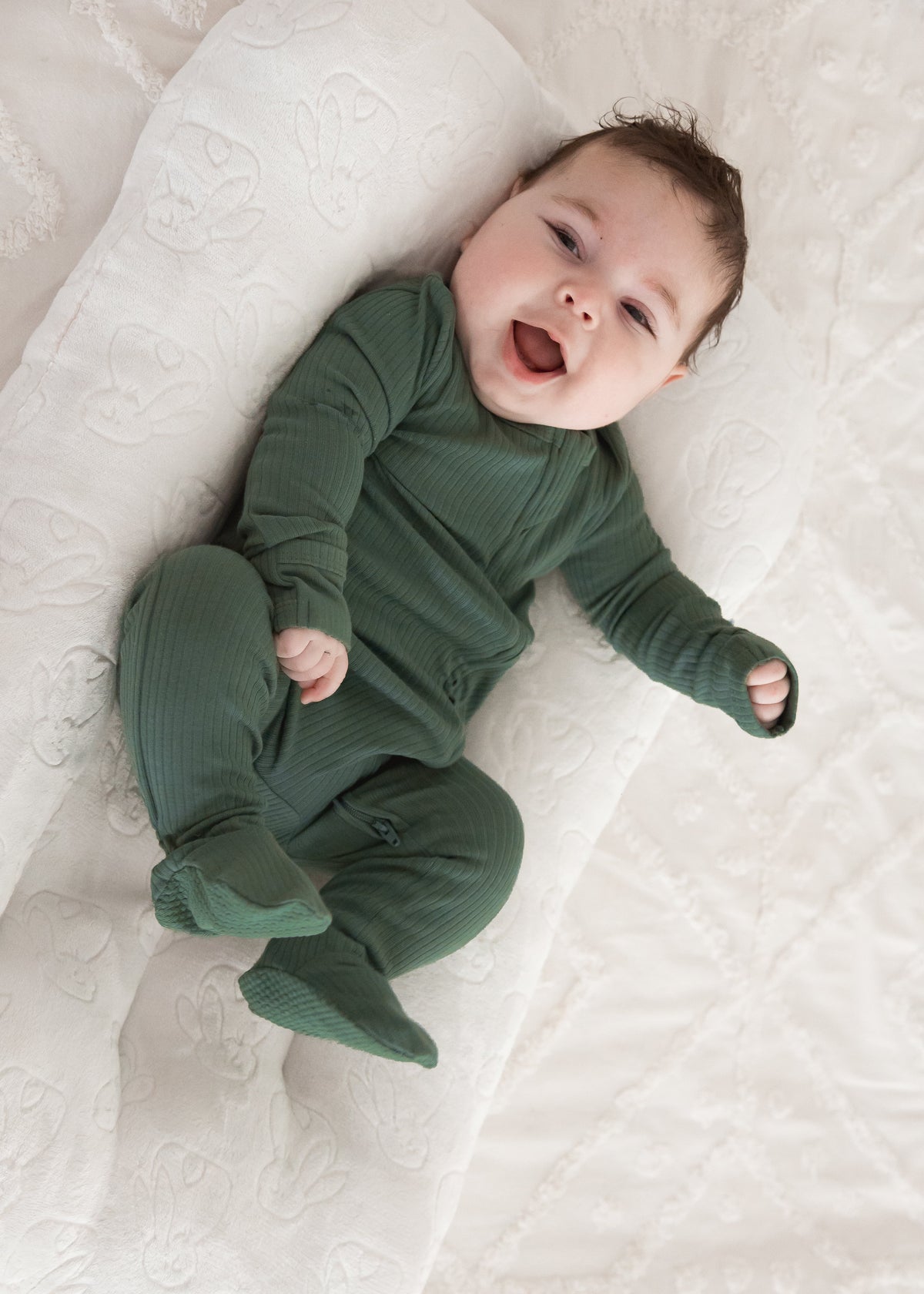 A happy baby smiling while lounging in a plush white baby nest, wearing the Pine green organic cotton ribbed sleeper.