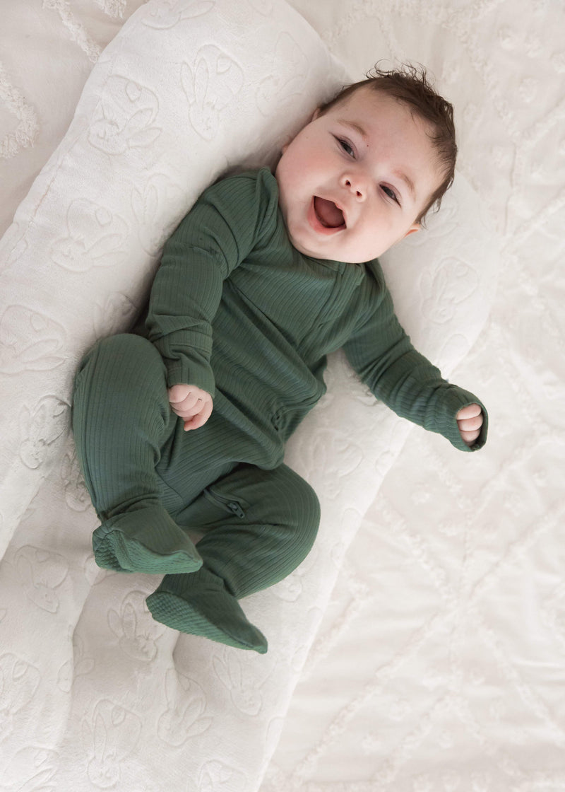 A happy baby smiling while lounging in a plush white baby nest, wearing the Pine green organic cotton ribbed sleeper.