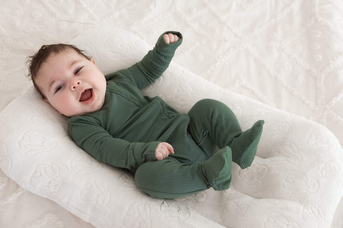 A smiling baby lying on their back on a white quilted lounger, wearing a dark green ribbed organic cotton footie from Milana Label.