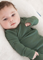 Full view of a baby looking directly at the camera while lounging in a white baby nest wearing the Pine green organic cotton footie sleeper.