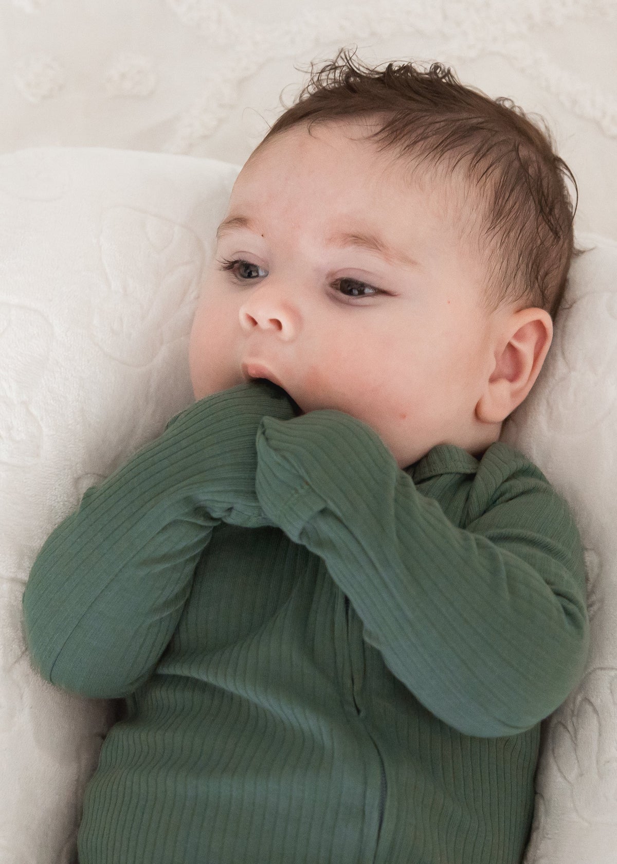A baby looking upwards while resting in a plush white lounger, wearing the Pine green ribbed baby sleeper with fold-over scratch mitts.