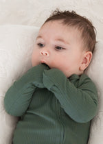 A baby looking upwards while resting in a plush white lounger, wearing the Pine green ribbed baby sleeper with fold-over scratch mitts.