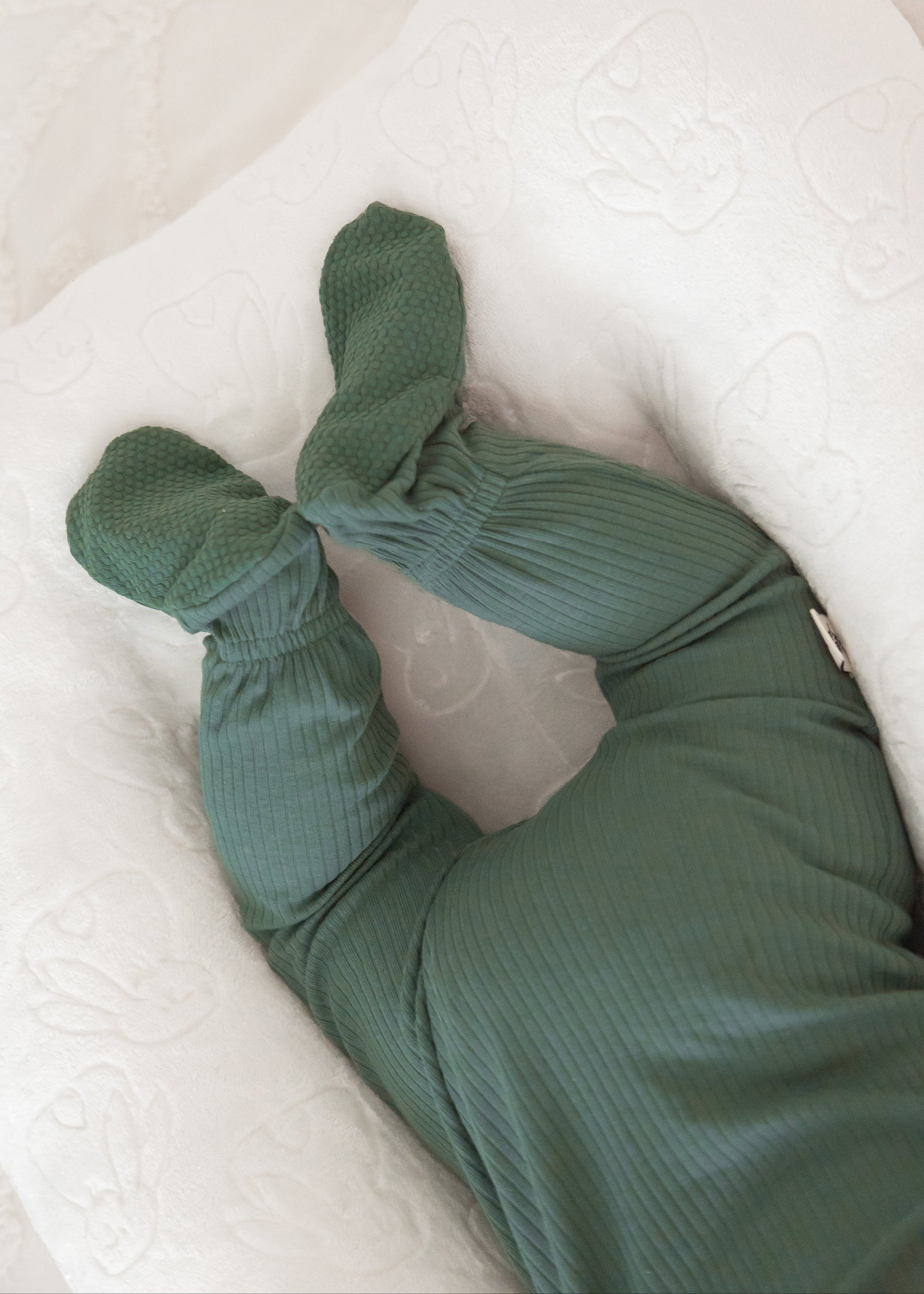 Close-up shot of a baby's legs and feet resting in a white baby nest, highlighting the rich texture of the Pine green ribbed organic cotton fabric.