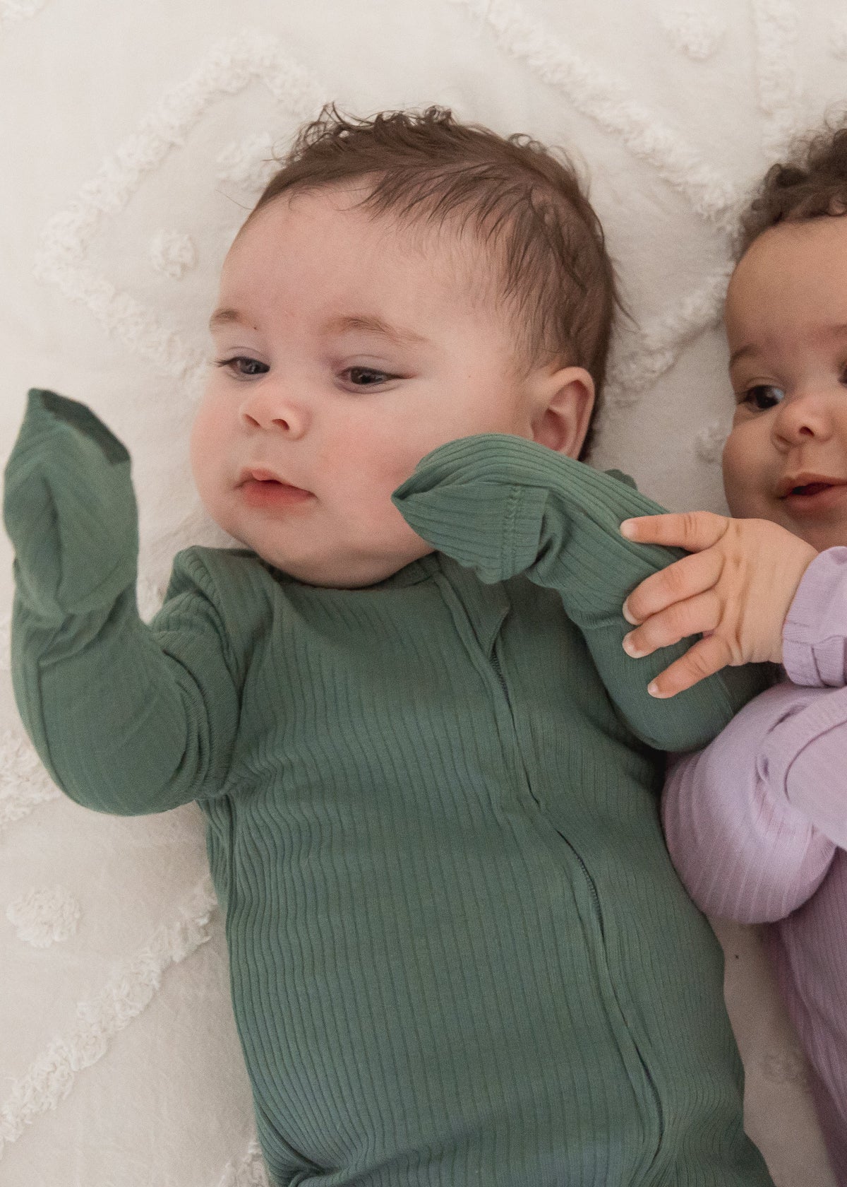 Close-up view of two babies interacting on a white blanket, one wearing the Pine green ribbed sleeper and the other in the Lavender Blush sleeper.