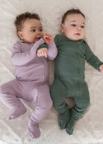 Two babies lying side-by-side on a white textured blanket, one wearing the Milana Label Lavender Blush ribbed footie sleeper and the other in their Pine sleeper.
