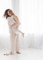 A woman and a young girl wearing matching pajamas with a small floral pattern are posed in a bright room with white sheer curtains and light wood flooring. The woman holds the girl in her arms, and a small matching jar sits on the floor nearby.