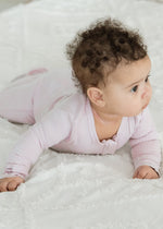 A baby enjoying tummy time on a textured white blanket, wearing the Milana Label organic cotton ribbed footie sleeper PJ in Petal pink.