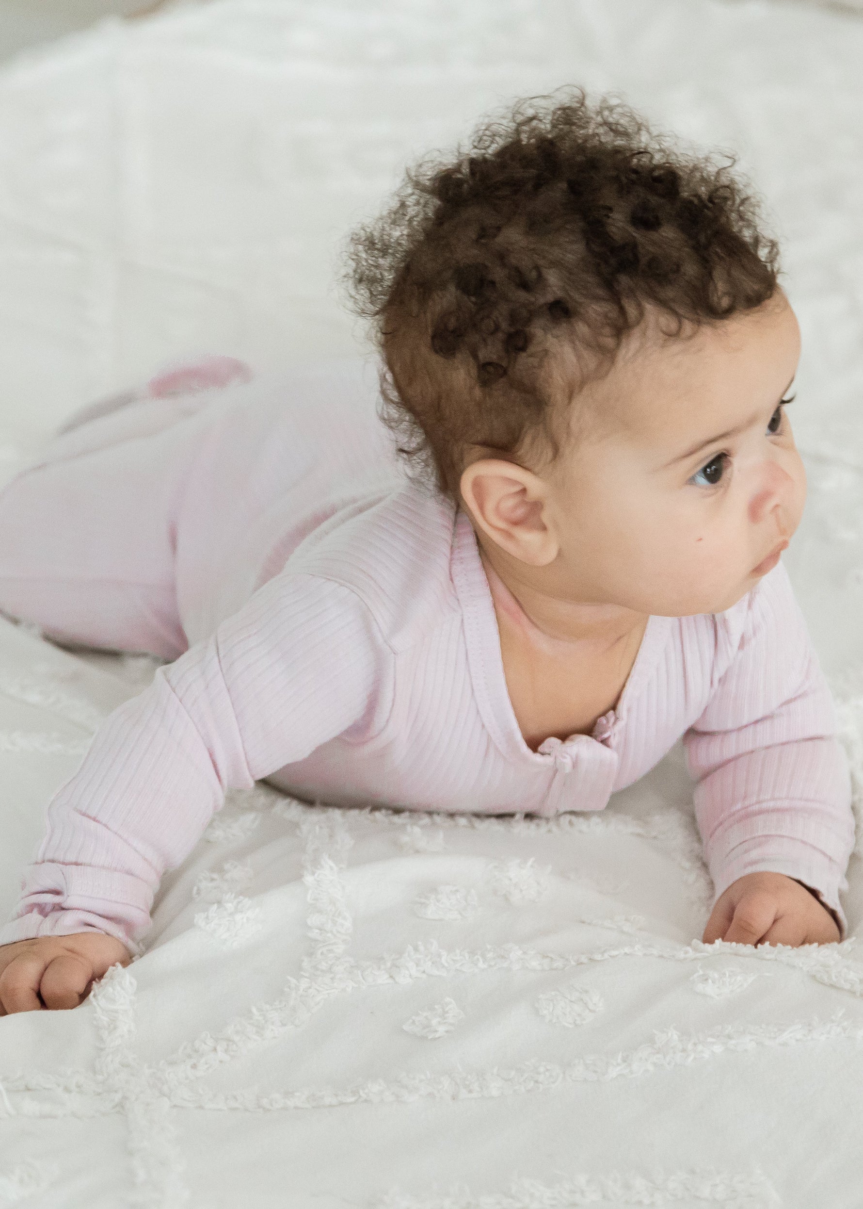A baby enjoying tummy time on a textured white blanket, wearing the Milana Label organic cotton ribbed footie sleeper PJ in Petal pink.