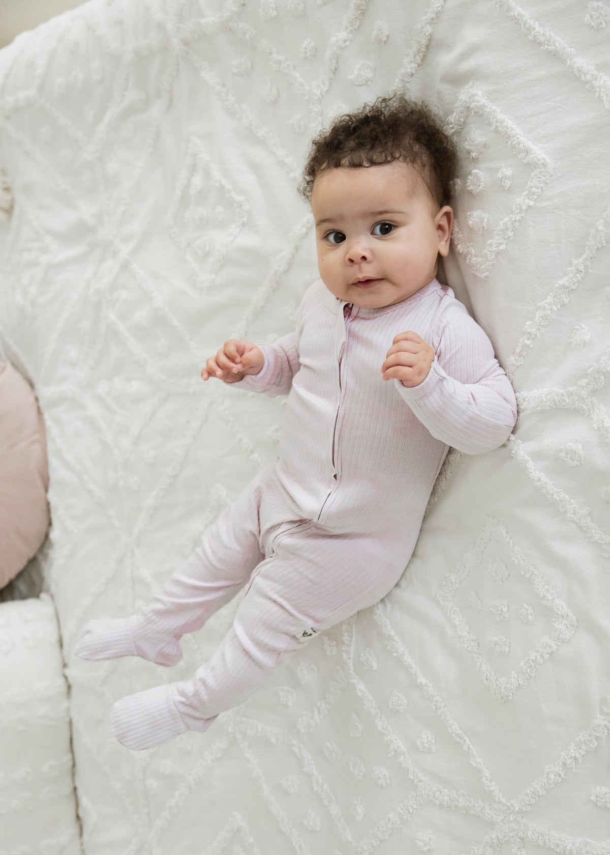 A baby girl wearing an organic cotton footie sleeper in the colour Petal pink by Milana Label lying on a neutral bedspread.
