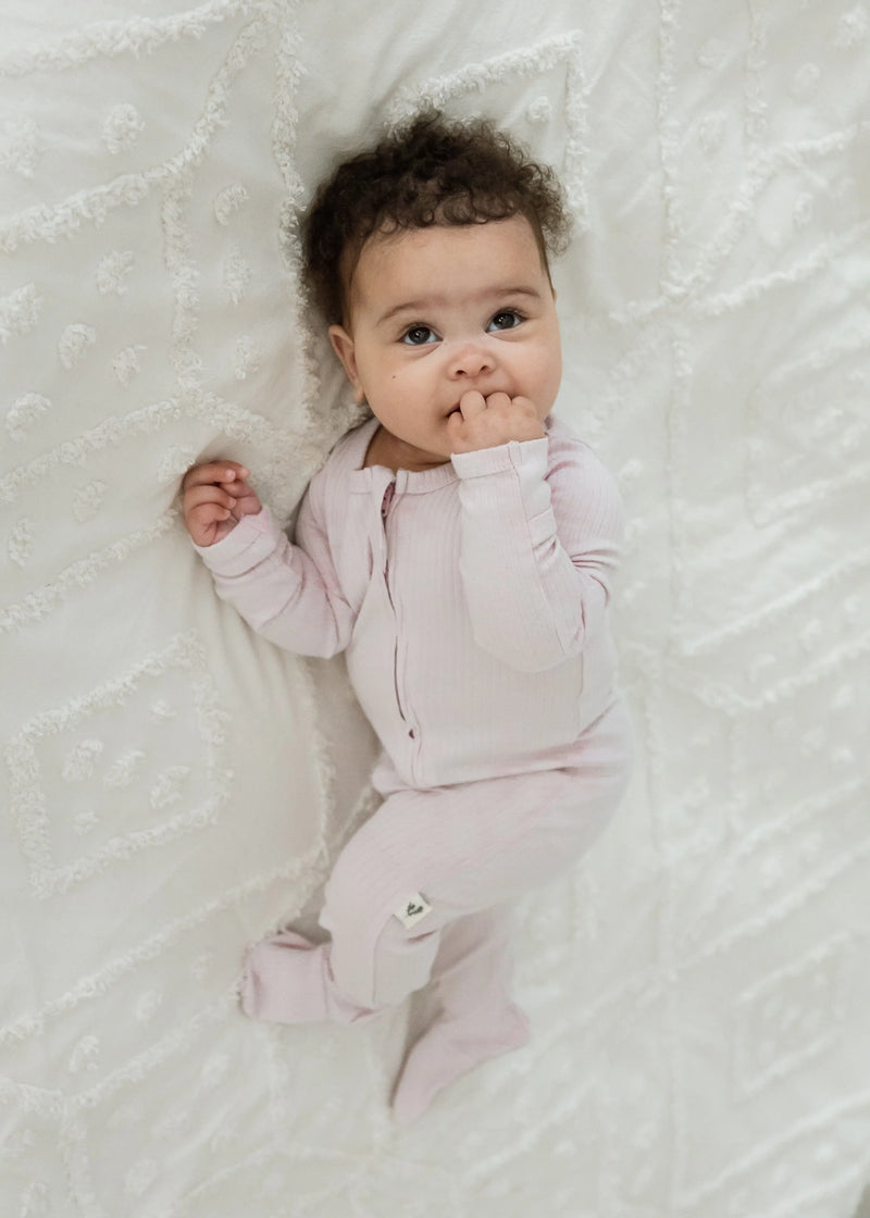 A baby looking directly up at the camera while resting comfortably on a white blanket in the Petal pink ribbed baby sleeper.