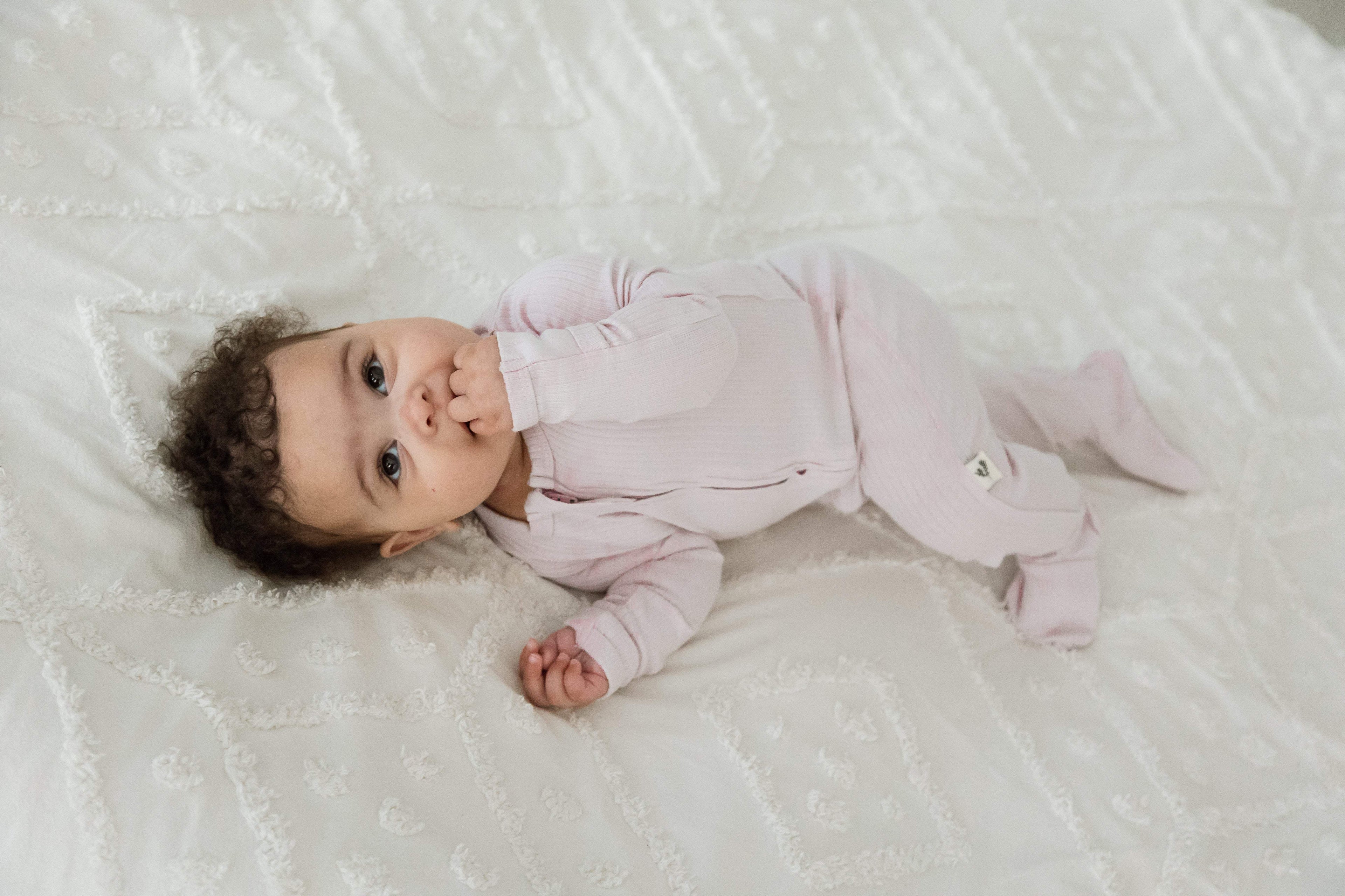 A baby lying on a white textured blanket, wearing a light pink long-sleeve footed onesie with double zipper and fold-over mitts.