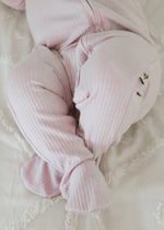 Close-up detail of a baby's legs and feet resting on a white blanket, showing the soft ribbed fabric of the Petal pink sleeper.