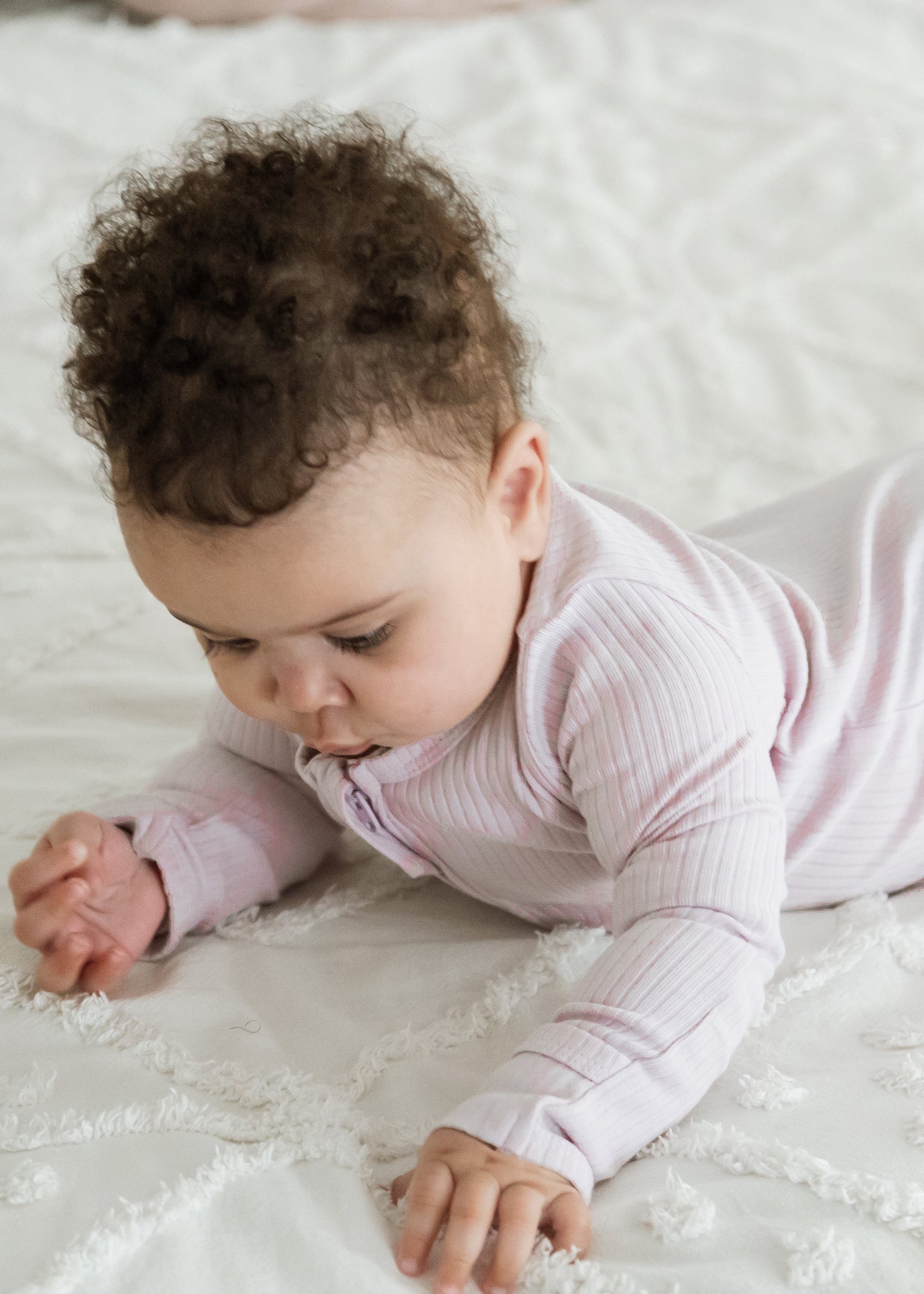 A baby lying on a textured white quilted blanket, wearing a light pink ribbed long-sleeve footed sleeper.