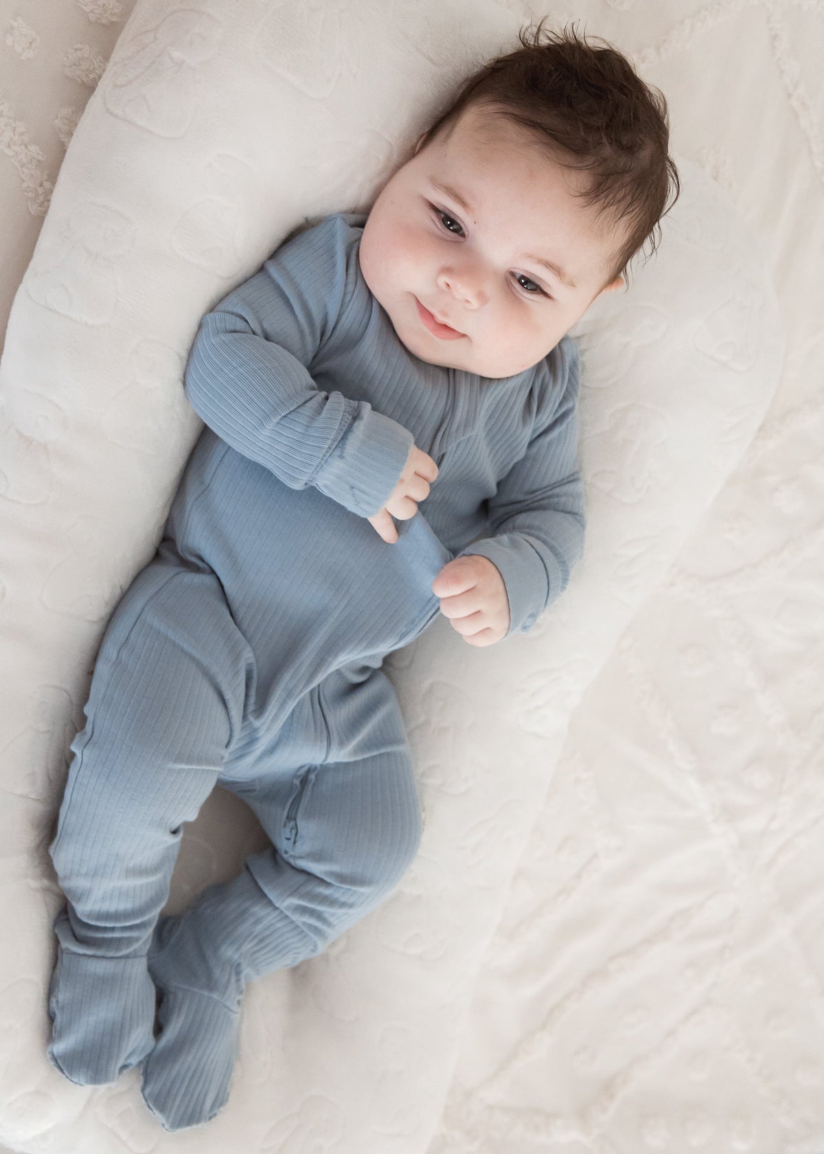 Full-length view of a baby resting happily in a plush white baby nest, wearing the Milana Label Soft Slate blue organic cotton ribbed footie sleeper.
