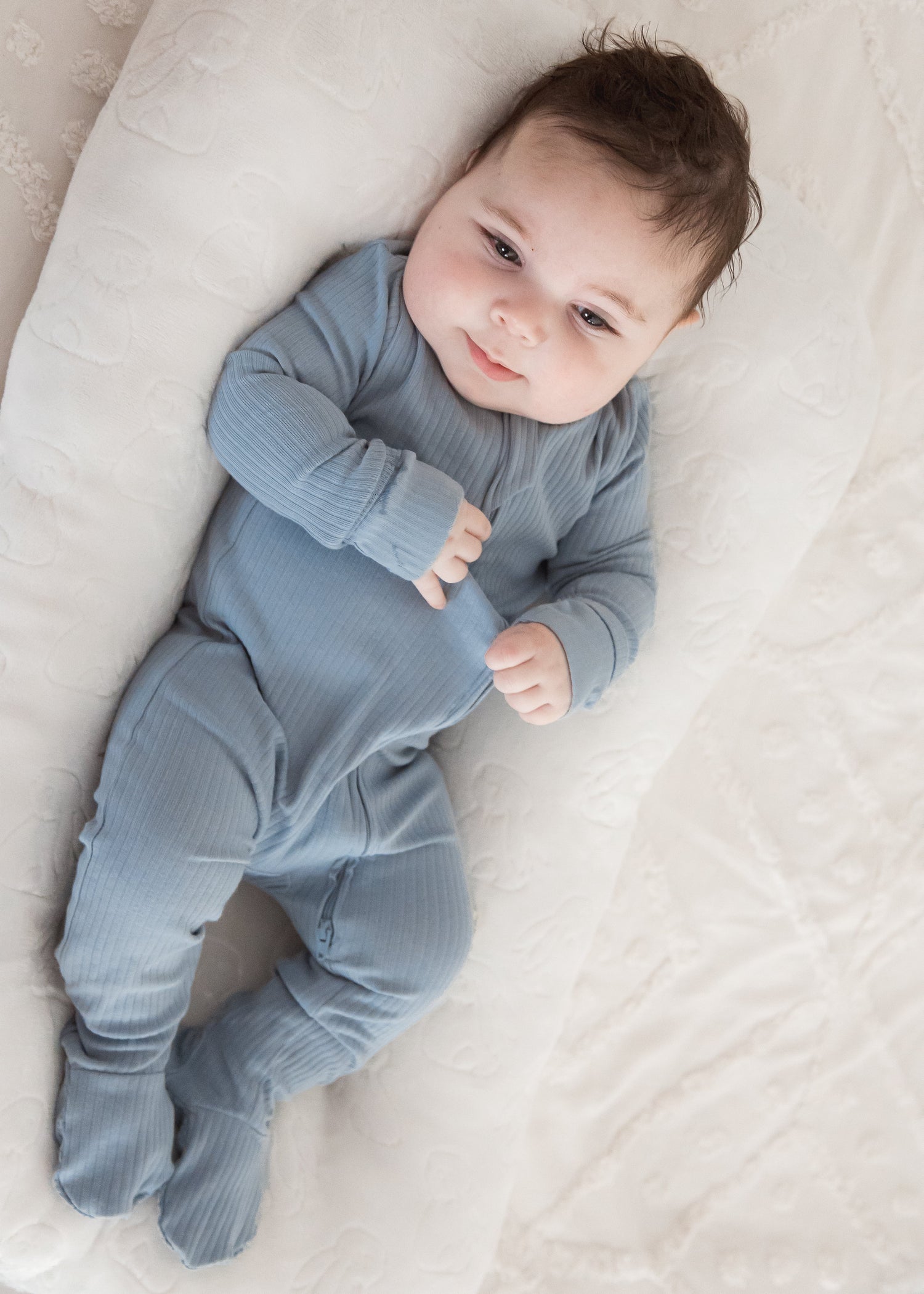 Full-length view of a baby resting happily in a plush white baby nest, wearing the Milana Label Soft Slate blue organic cotton ribbed footie sleeper.