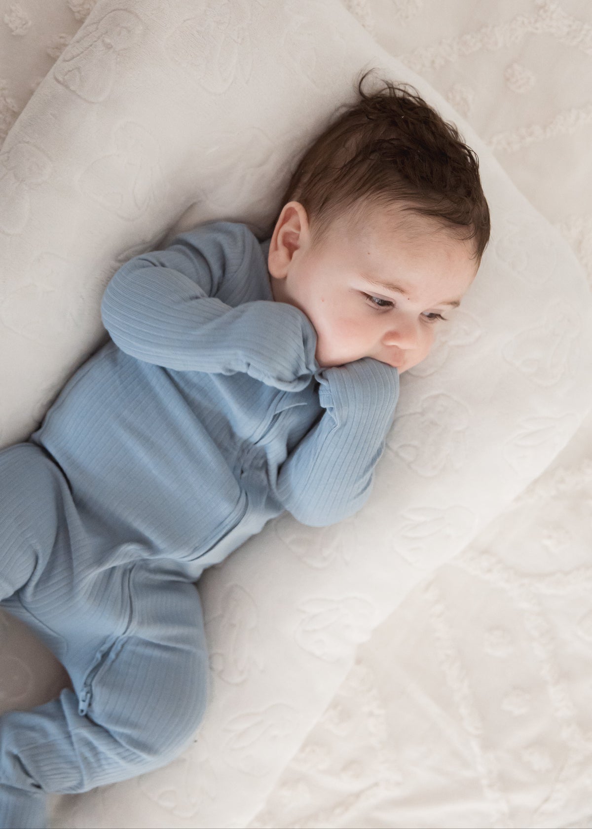 A baby looking off to the side while resting in a cream-colored lounger, wearing the Soft Slate blue ribbed baby sleeper.