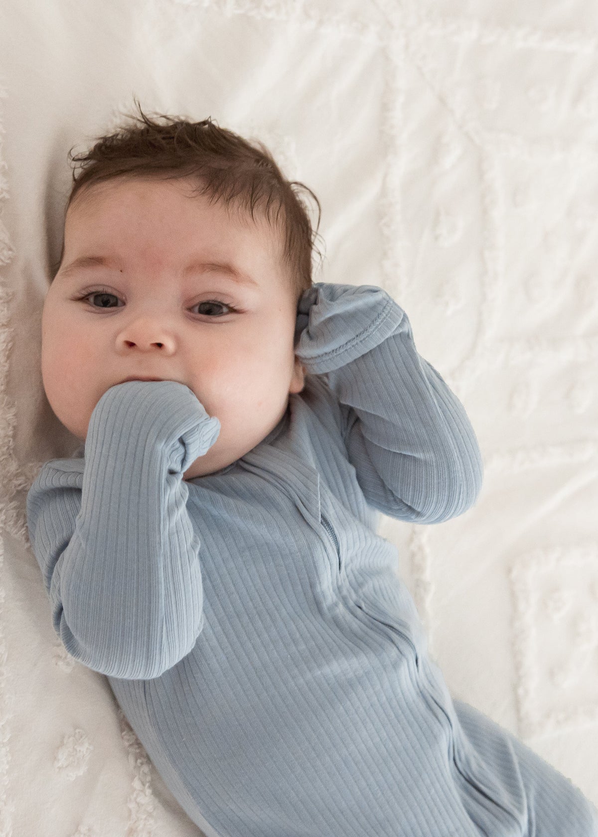 Close-up view of a baby looking up at the camera while wearing the Soft Slate blue organic cotton sleeper, with fold-over mitts covering their hands.