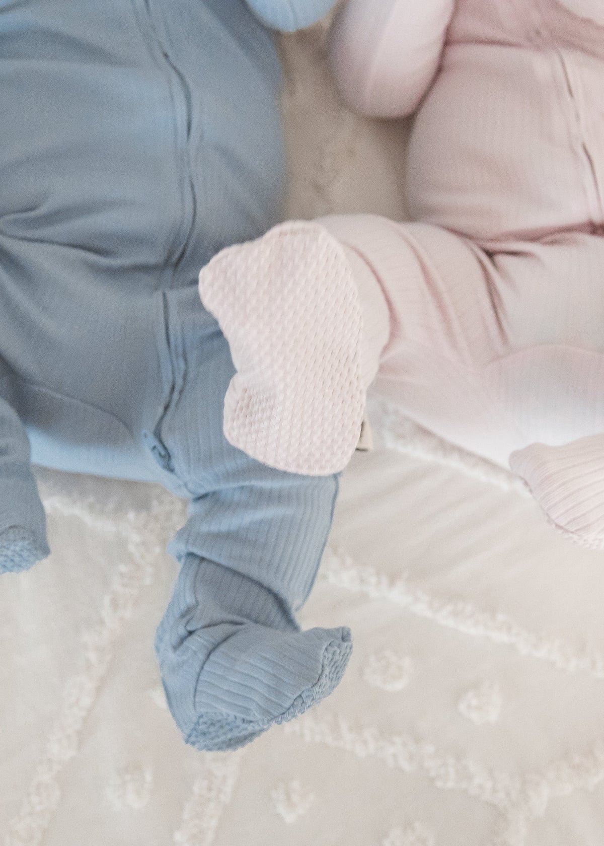 Lower angle view of two babies lying together on a textured blanket, showcasing the feet and fabric of the Soft Slate blue and Petal pink ribbed sleepers.