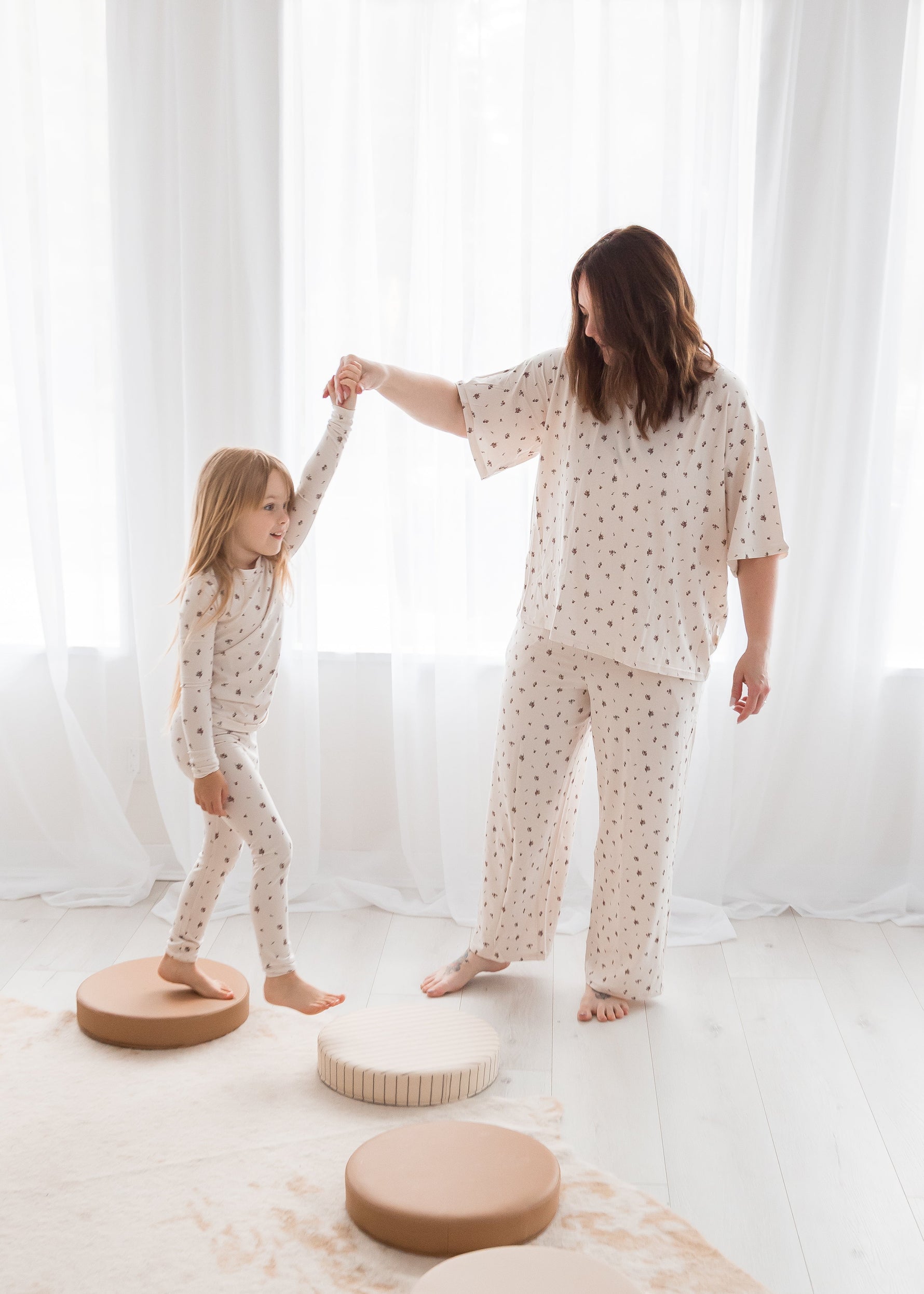 A mother and daughter wearing matching light-colored patterned pajamas, holding hands as the young girl balances on circular stepping cushions in a bright, sunlit room with sheer white curtains.