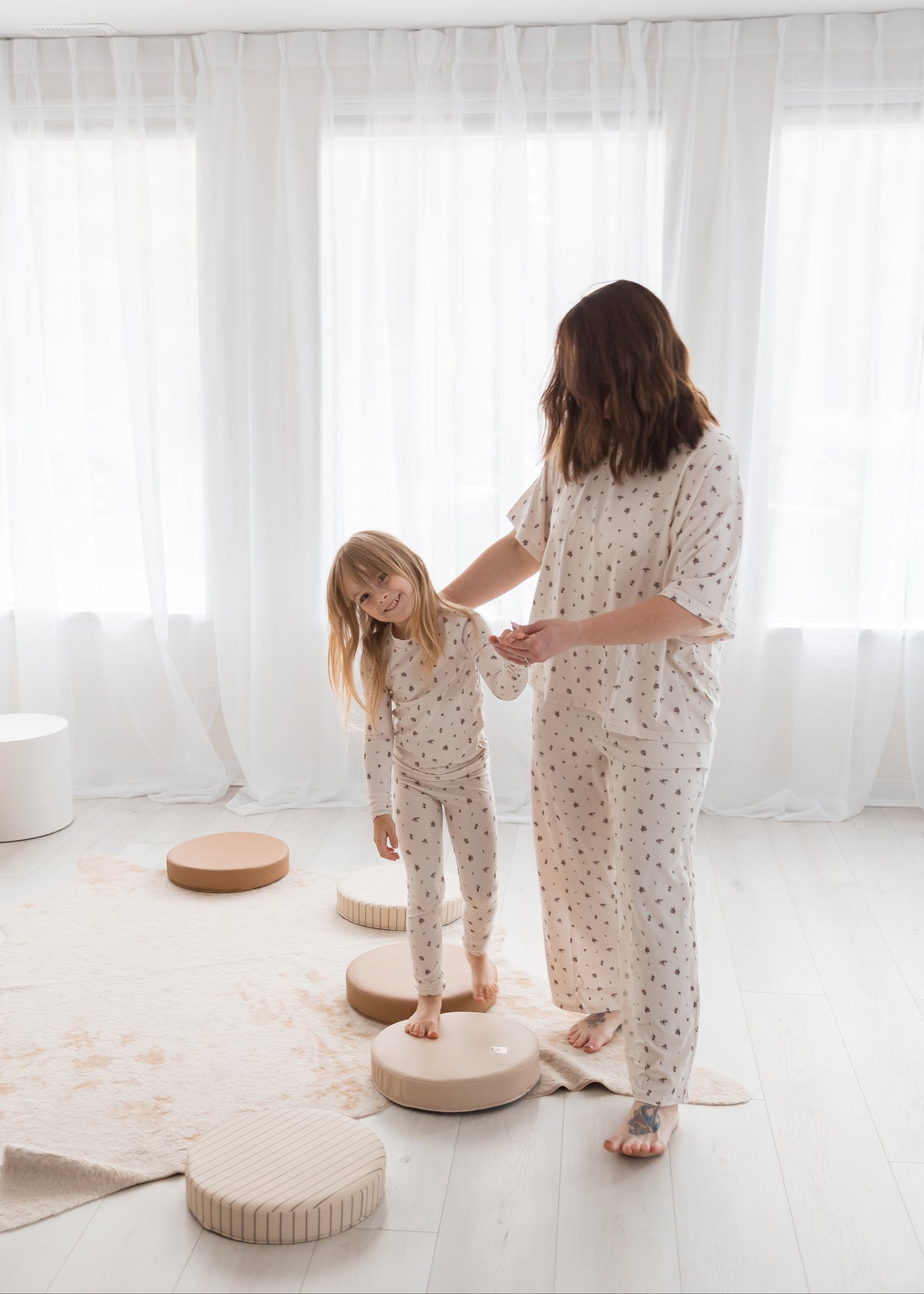 A mom holds her daughter's hand as the young girl steps across neutral-toned circular floor cushions. Both are wearing matching birch floral Tencel pajamas in a minimalist, bright living space.