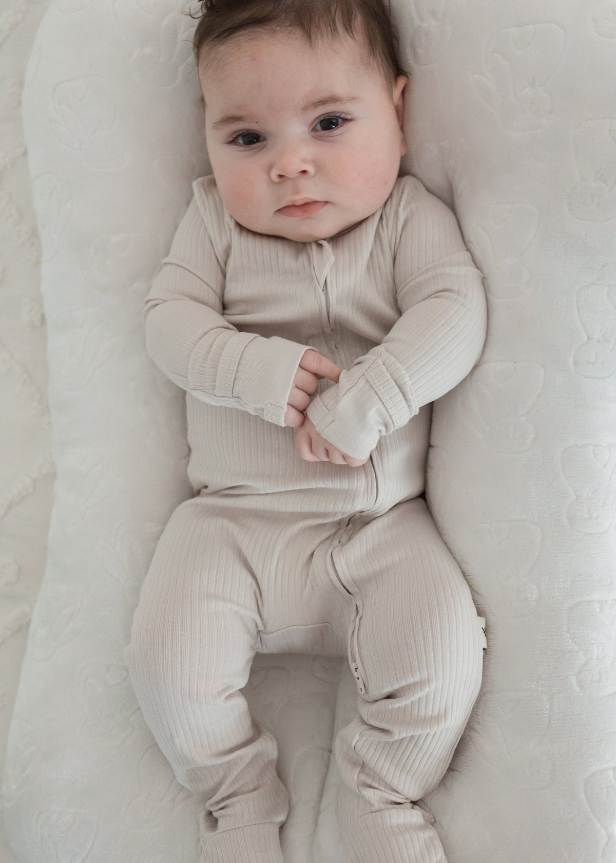 Full-length view of a baby looking at the camera while wearing the Milana Label Stone greige ribbed footie sleeper, lounging in a plush white baby nest.