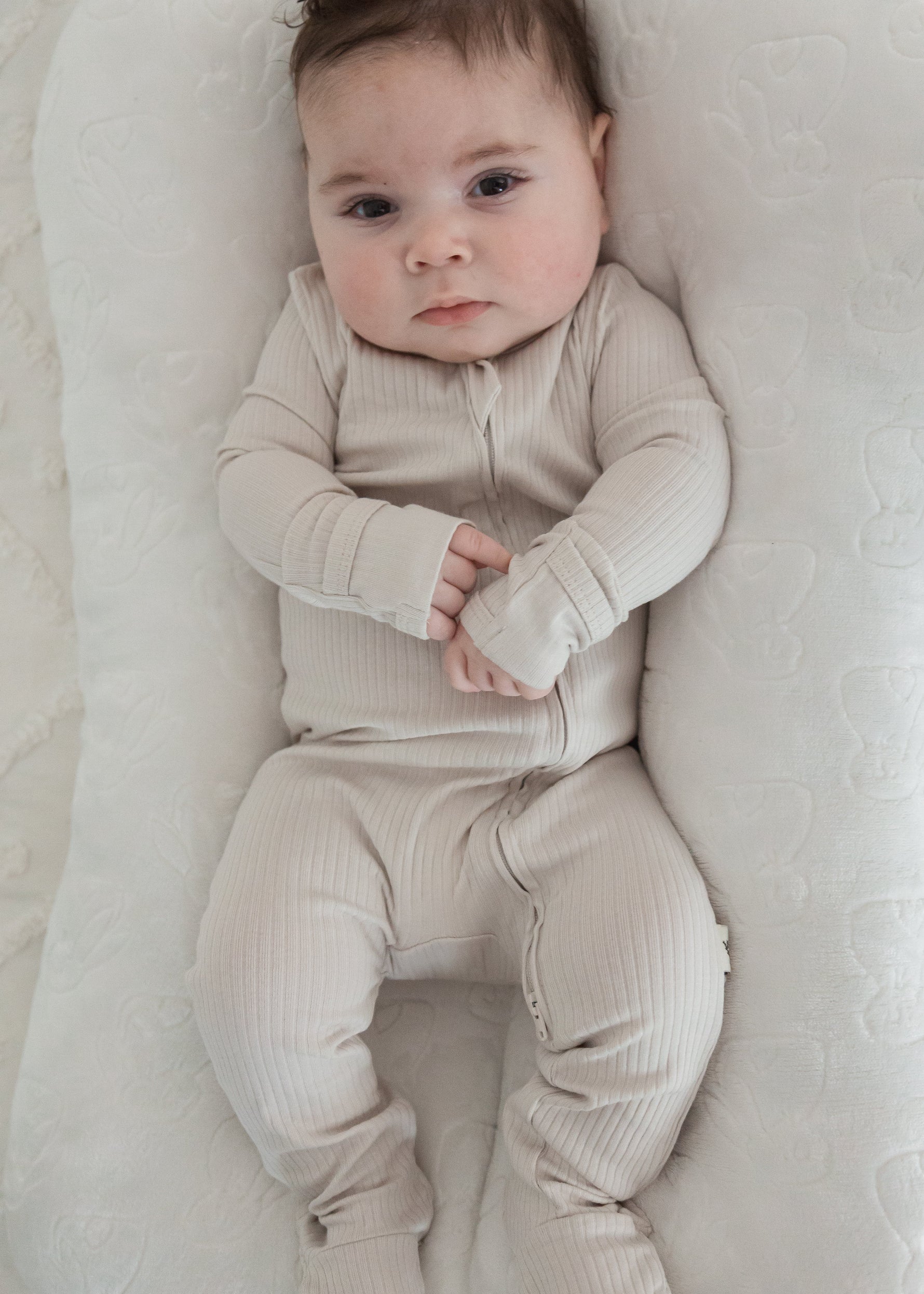 Full-length view of a baby looking at the camera while wearing the Milana Label Stone greige ribbed footie sleeper, lounging in a plush white baby nest.