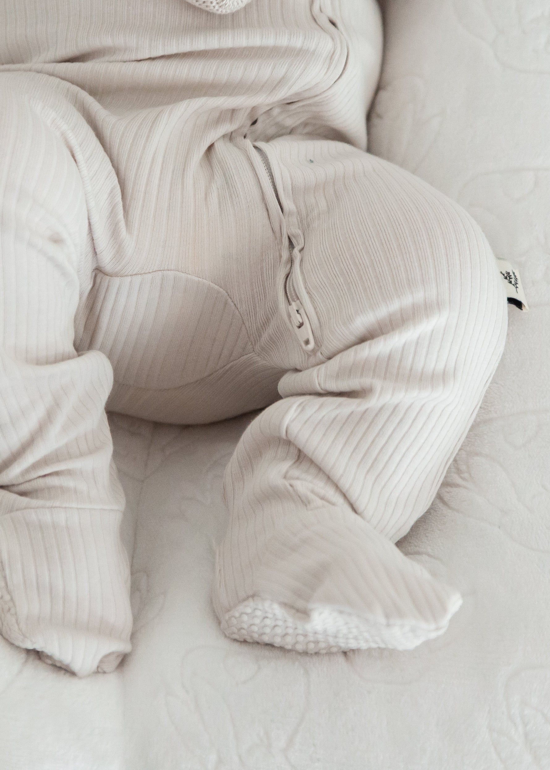 Close-up shot of a baby's legs and feet resting comfortably in a white lounger while wearing the Stone greige ribbed footie pajama.
