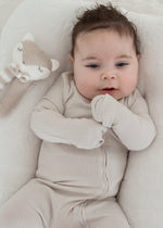 Full view of a baby resting in a cream-colored lounger wearing the Stone greige organic cotton ribbed footie sleeper.
