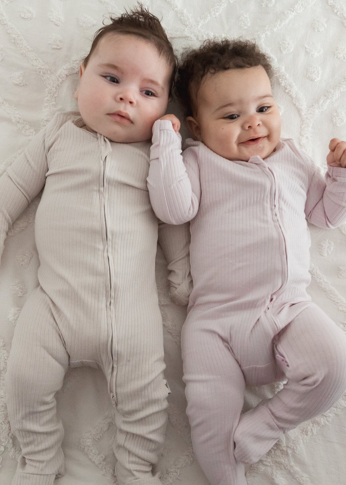 Two babies lying side-by-side on a white blanket, one wearing the Stone greige ribbed sleeper and the other in the Petal pink sleeper.