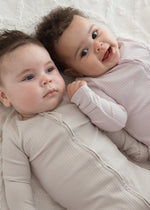 Two babies lying side-by-side on a white blanket, one wearing the Stone greige ribbed sleeper and the other in the Petal pink sleeper.