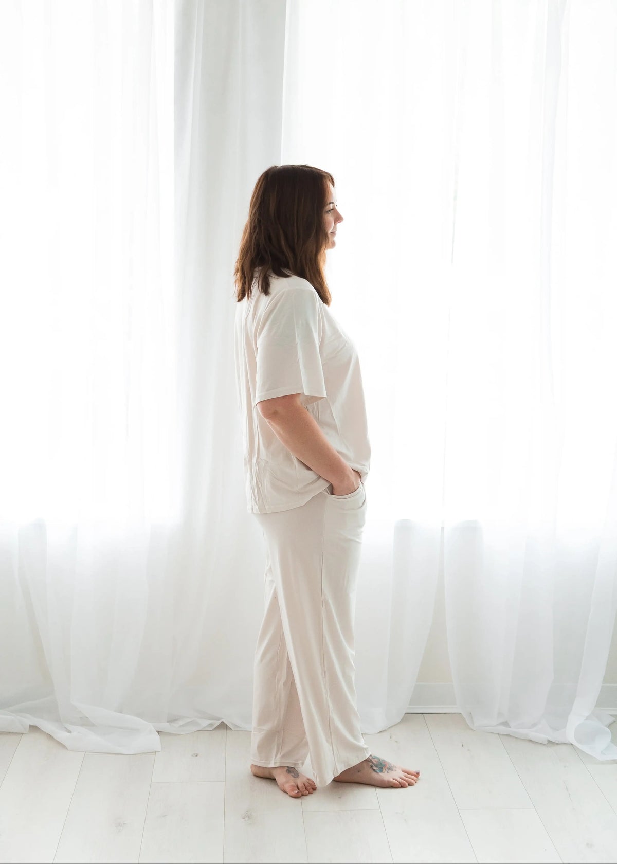 Full-length profile shot of a woman standing relaxed against a bright white window curtain backdrop, showcasing the drape and comfortable fit of a light birch-colored two-piece pajama set.