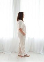 Full-length profile shot of a woman standing relaxed against a bright white window curtain backdrop, showcasing the drape and comfortable fit of a light birch-colored two-piece pajama set.